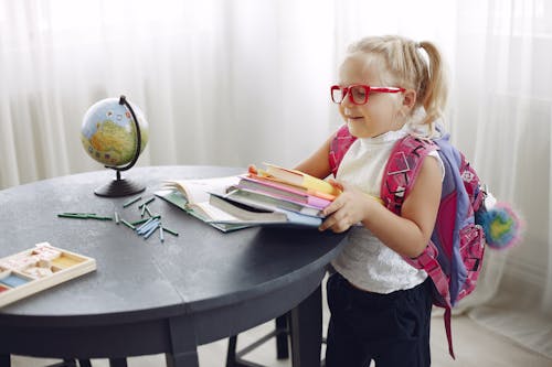 Little girl wearing casual clothes and glasses smiling while preparing books for homework at home during weekend and looking away