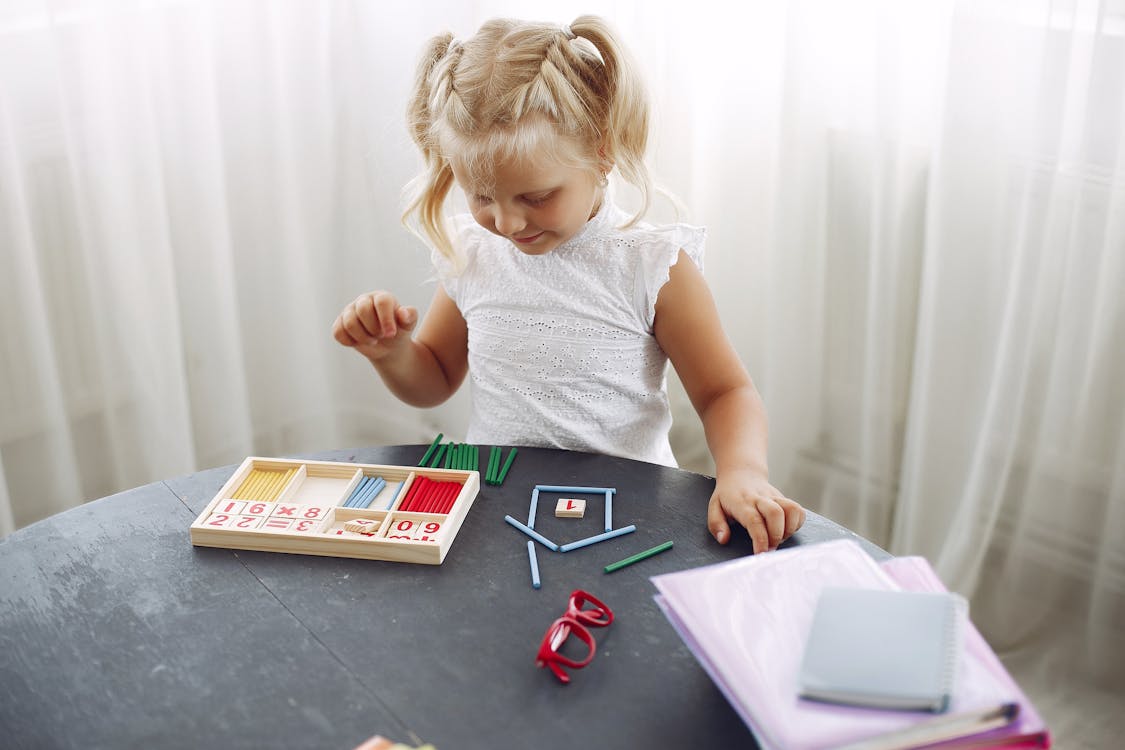Free Blond little girl standing while playing with puzzle in living room at home and counting sticks different colors during weekend Stock Photo