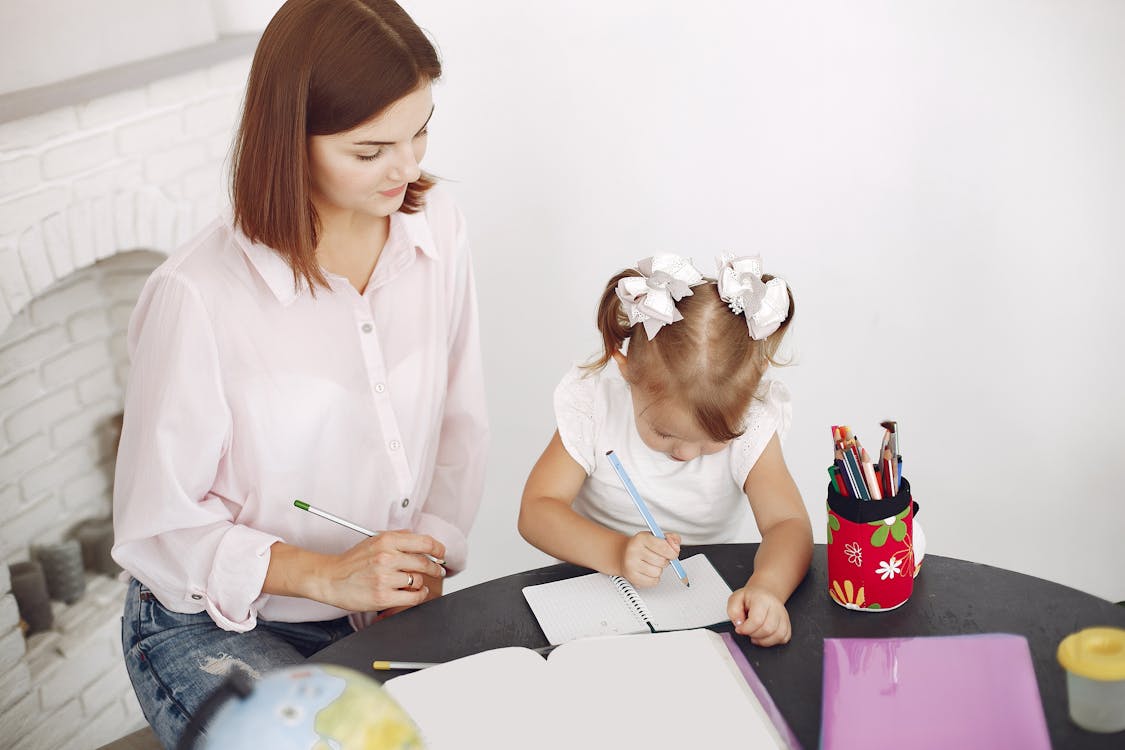 Free From above small girl drawing with pencil in notepad while mother sitting near at round table Stock Photo