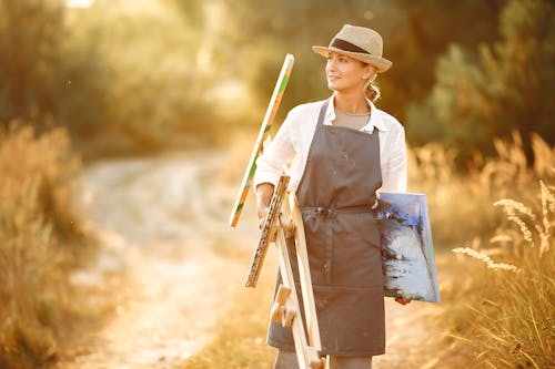 Happy female in hat and apron carrying easel and canvas while walking on road in summer sunny day in nature