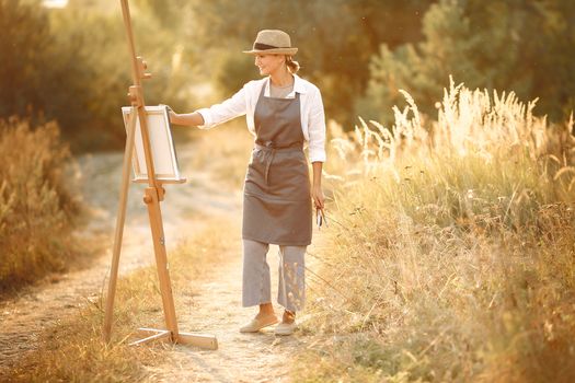 Full body of cheerful female wearing hat and apron standing on road in nature and drawing pictures with brush while spending time in countryside