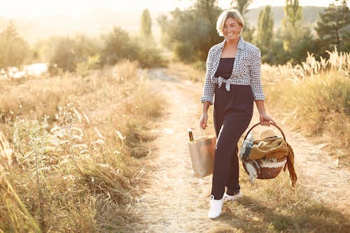 Full body of excited blond woman smiling and looking at camera while walking on path with basket and resting during weekend