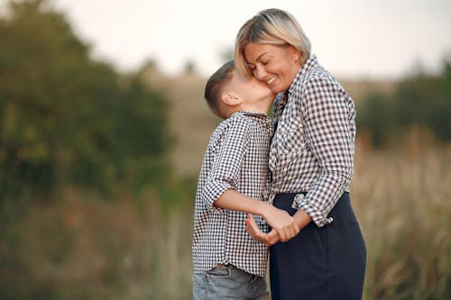 Side view of cheerful middle aged woman in plaid shirt standing with grandson while receiving kiss on cheek from boy on blurred background of field in summer