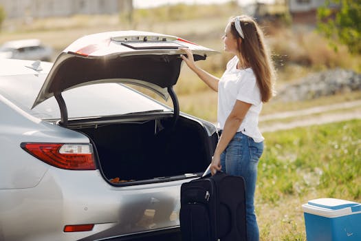 Side view of cheerful female traveler in headband and casual clothes putting luggage in open trunk of modern car while spending summer weekend in countryside