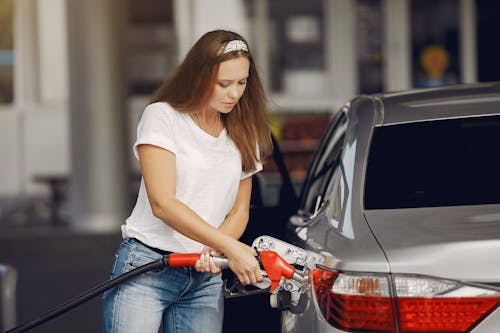 Attentive female driver in casual outfit and headband filling up modern automobile with automotive fuel gun on petrol station while looking down