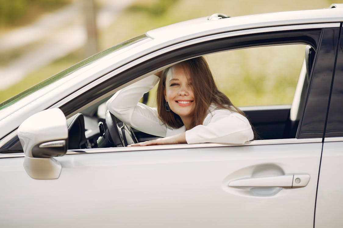 Cheerful female driver in white shirt sitting inside modern automobile and looking at camera through opened side window with smile during car trip in summer day