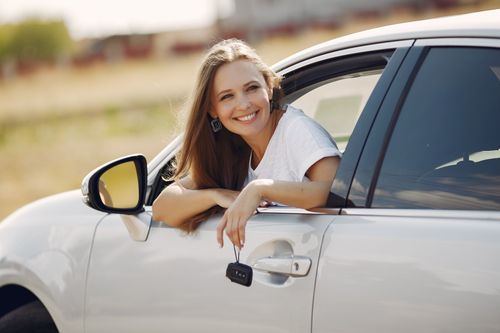 Free Happy female driver in white t shirt leaning out of side door window with car key in hand and looking away with smile in sunny summer day Stock Photo