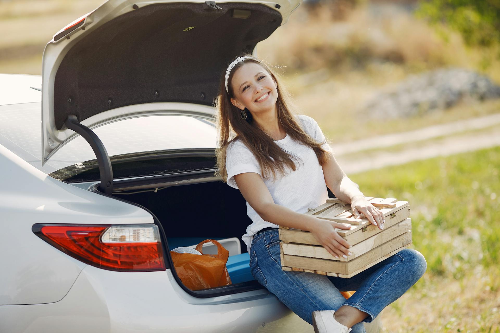 Cheerful female traveler in headband and casual clothes sitting in open trunk of car with wooden box while spending summer weekend in countryside and smiling looking away
