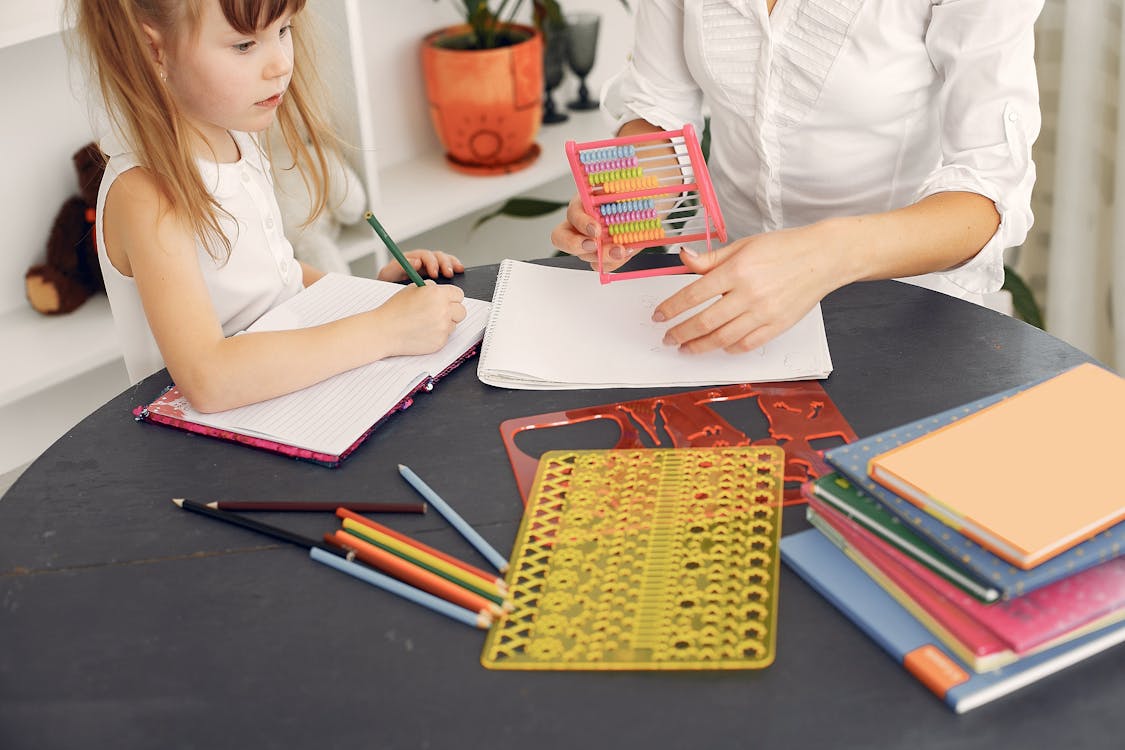 From above of little girl sitting at table with school items and textbooks while writing in notebook with pencil together with unrecognizable young home teacher in white shirt with abacus in hand during early home education