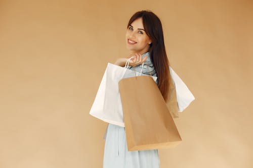 Side view of happy female shopper carrying paper bags while looking at camera over shoulder and standing isolated on brown background