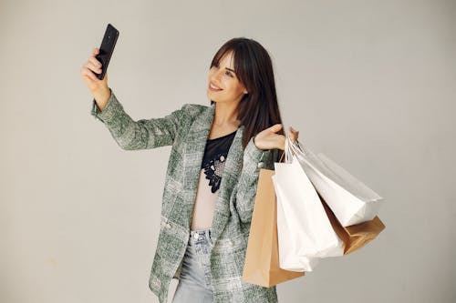 Stylish female shopper in trendy plaid jacket and jeans standing against gray background with paper bags in raised arm while using smartphone for photo