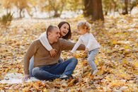 Laughing adult parents in warm clothes sitting together on ground in autumn park while catching adorable little daughter with curly hair running on fallen autumn maple leaves