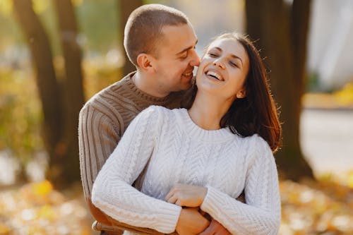 Tender adult man and laughing young woman hugging with closed eyes while standing close to each other during romantic date in park