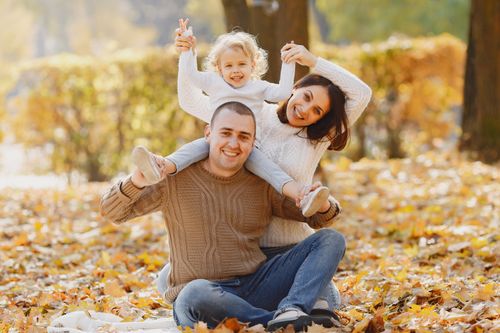 Free Smiling young parents in warm clothes sitting together on lawn in autumn park on white napkin while spending time together with cute little daughter sitting on shoulders of father and looking at camera Stock Photo
