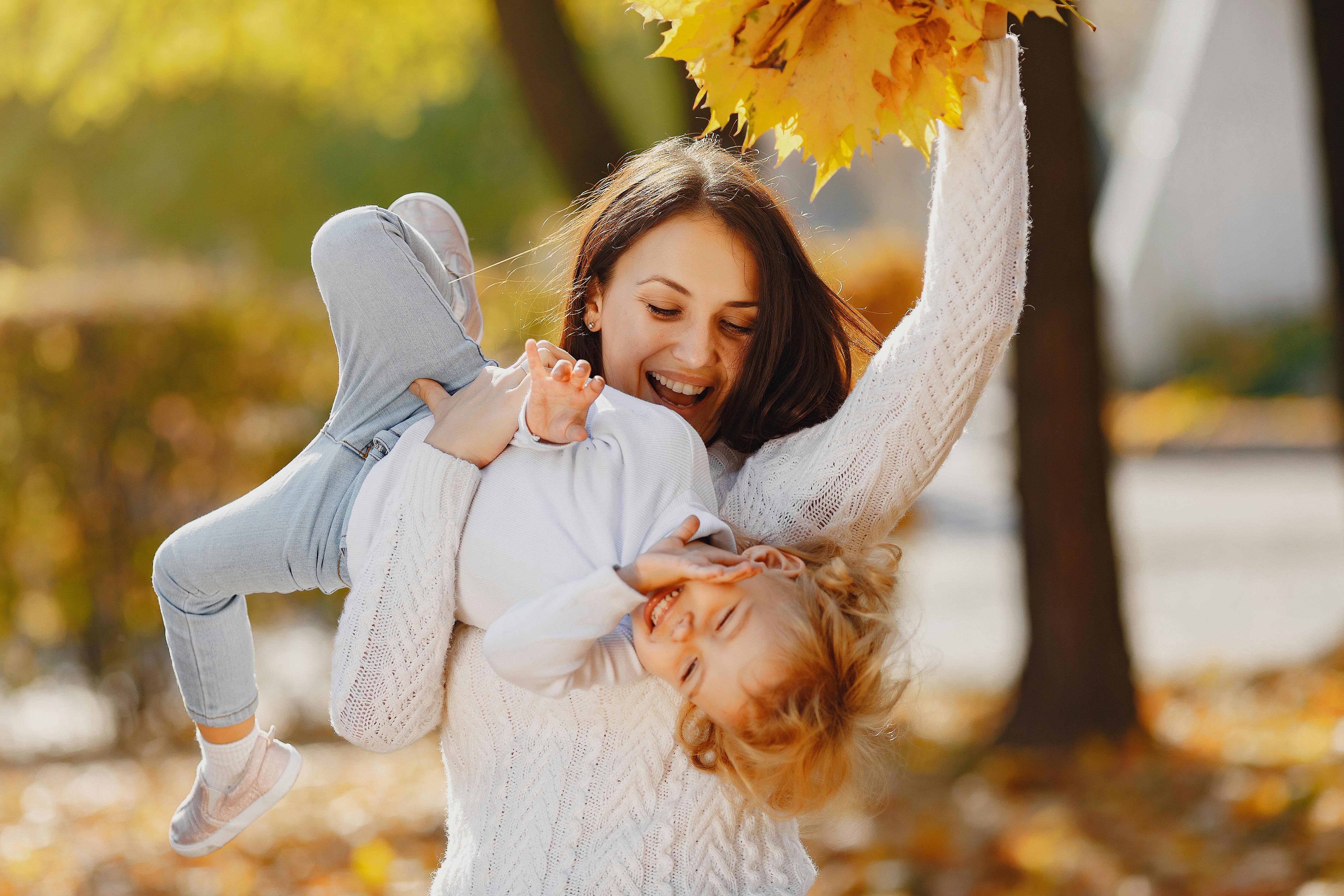 Happy young woman in knitted sweater carrying little girl and playing while showing bunch of yellow leaves in hand on top during walk in autumn park