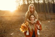 Cheerful young woman in casual outerwear with bunch of yellow leaves carrying on shoulders daughter while walking in autumn park on sunset