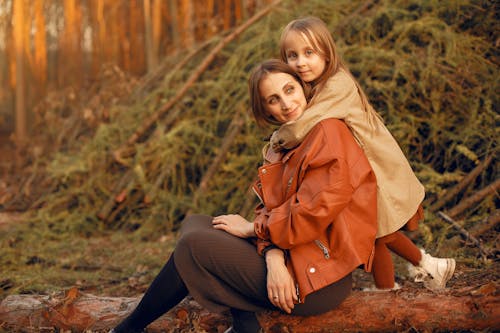 Free Side view of smiling stylish mother and adorable daughter wearing trendy outerwear sitting on log and hugging in autumn forest in evening sun Stock Photo