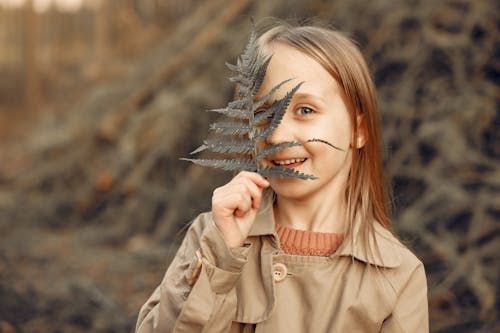 Happy little blond girl in stylish beige trench coat standing in autumn forest and looking at camera through green fern leaf and smiling