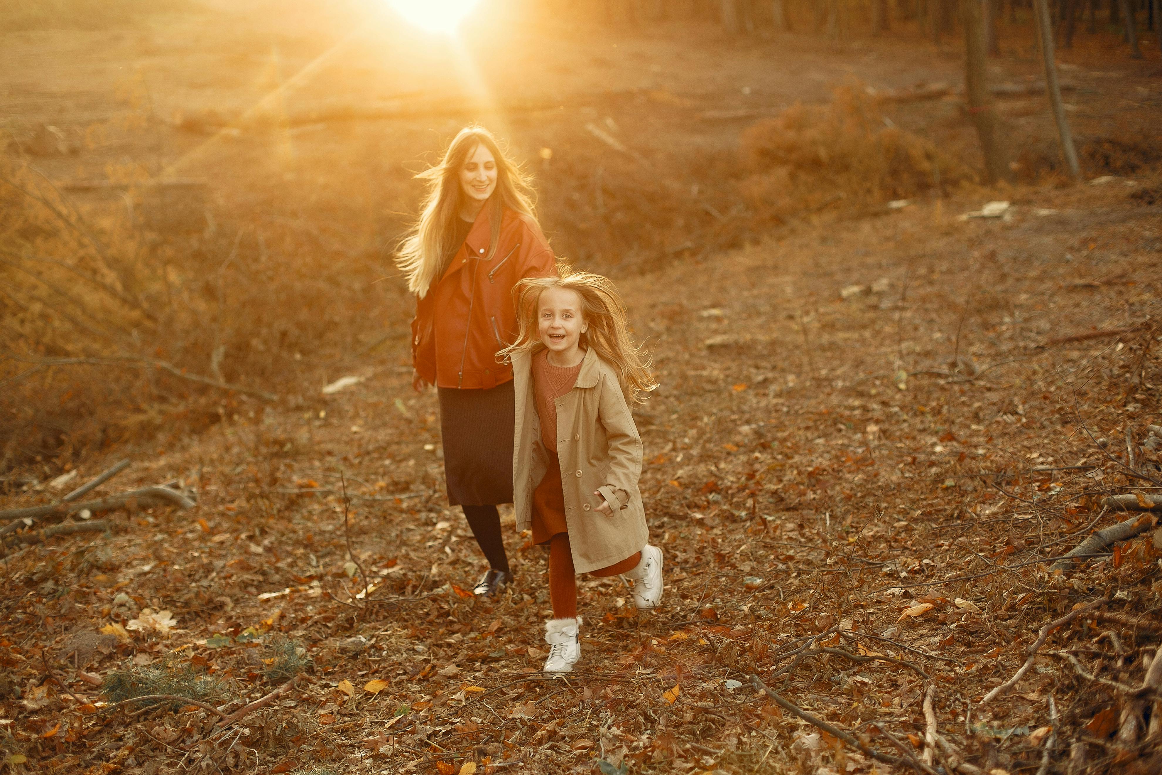 Full length of happy delighted blond mother and daughter in stylish outerwear smiling and walking together in autumn woods during sunset in back lit