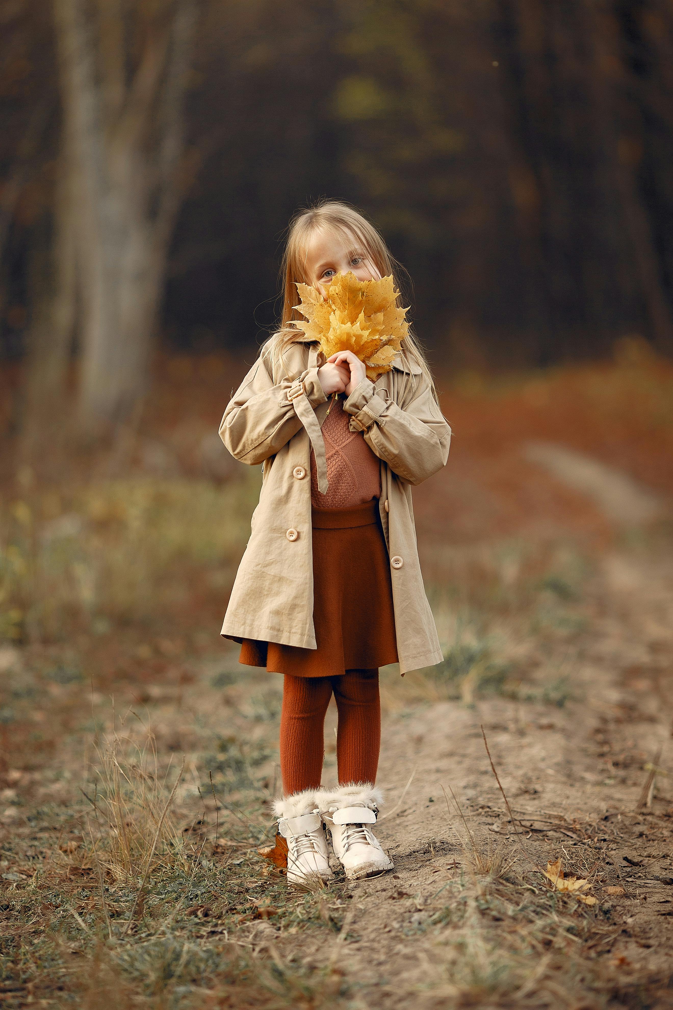 Full body of cute trendy girl in brown skirt and beige trench coat covering half of face with bunch of yellow autumn maple leaves in forest