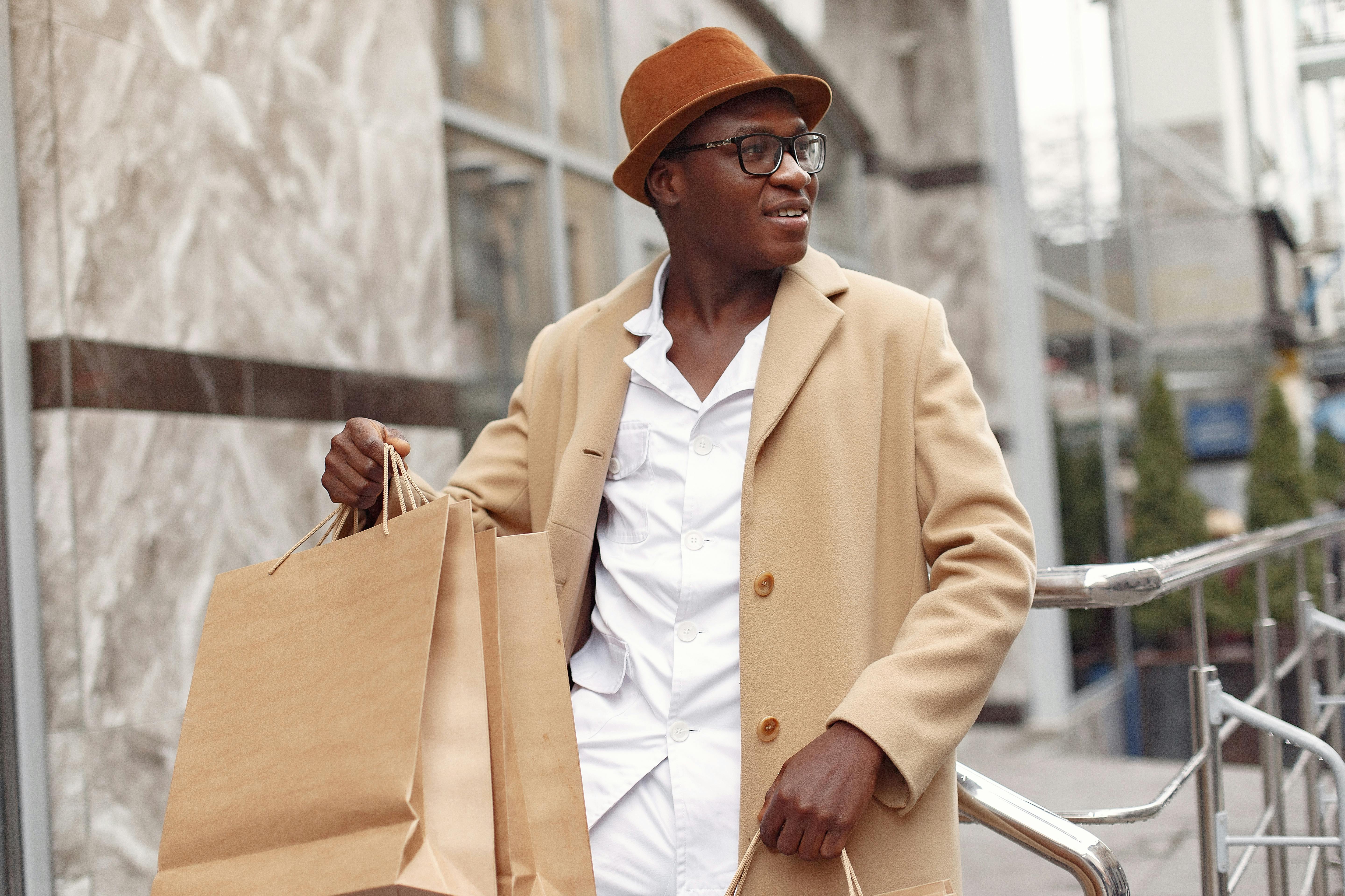 Confident stylish African American male in trendy beige coat and hat with paper shopping bags leaning on building railing in daytime and looking away happily