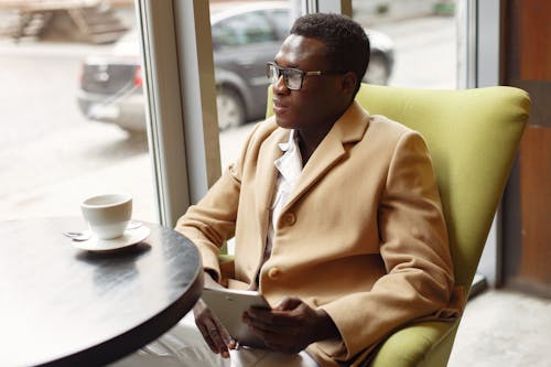 Free Serious African American male in trendy formal suit and eyeglasses sitting on cozy chair in cafe with cup of coffee and browsing tablet Stock Photo