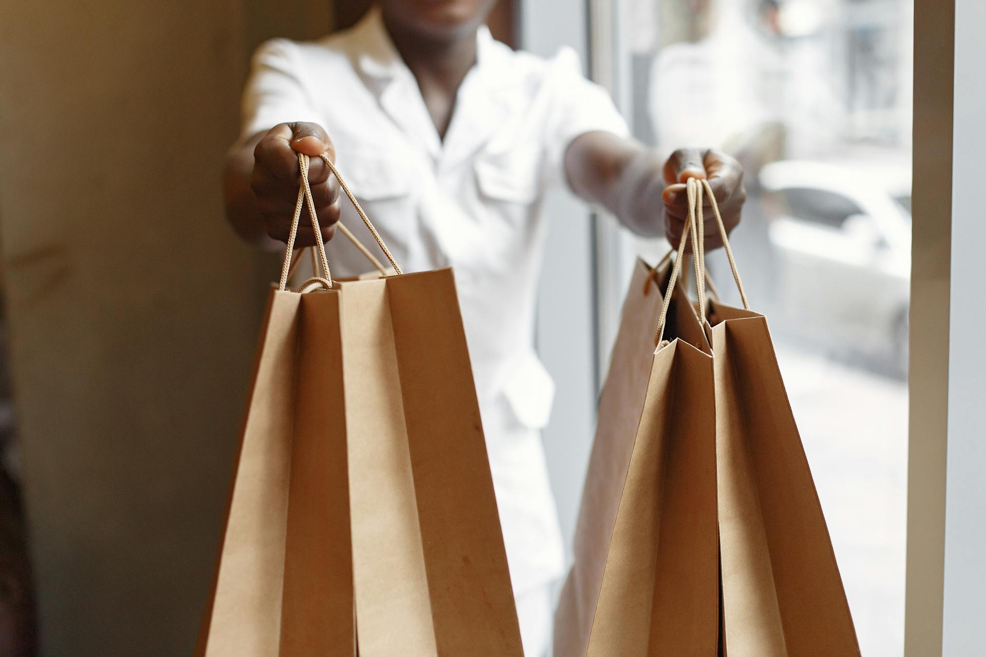 Crop anonymous African American person in white jacket passing purchases in paper shopping bags while standing near window in daylight