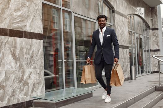 Full body of happy classy black male in elegant black suit carrying paper shopping bags while strolling along modern luxury marble wall building