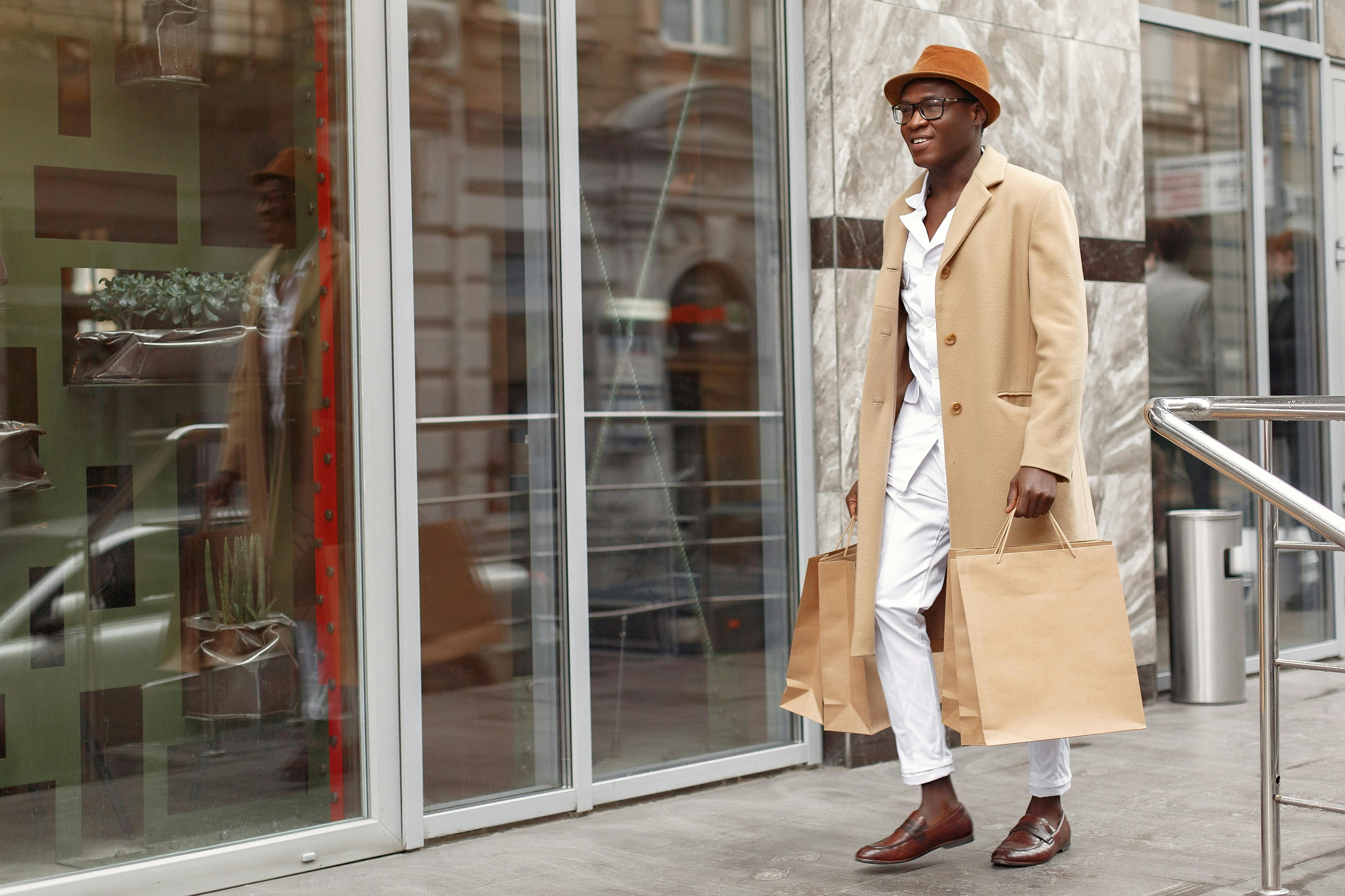 Full length of cheerful trendy African American male in white suit and beige coat walking with paper shopping bags against glass wall building