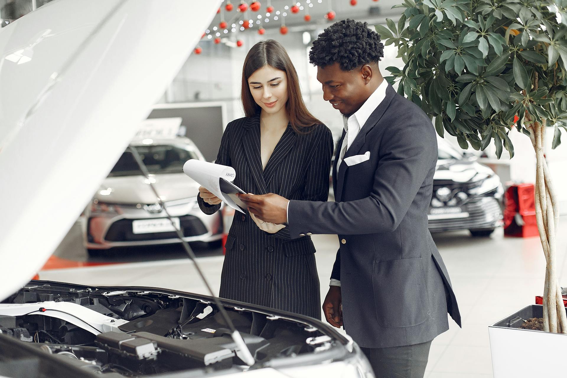 Man and woman reviewing documents at a car dealership next to an open car hood.