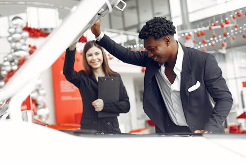 Positive smiling diverse stylish man and female dealer checking under car motor hood and smiling while choosing new car in car showroom
