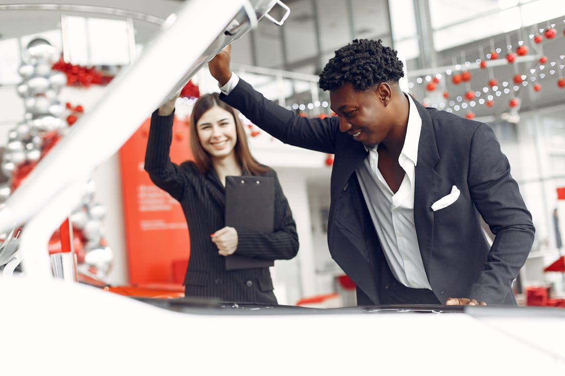 Free Positive smiling diverse stylish man and female dealer checking under car motor hood and smiling while choosing new car in car showroom Stock Photo