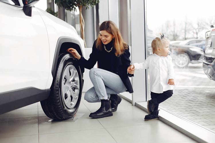 Happy Woman With Daughter Checking Car Wheel In Car Showroom