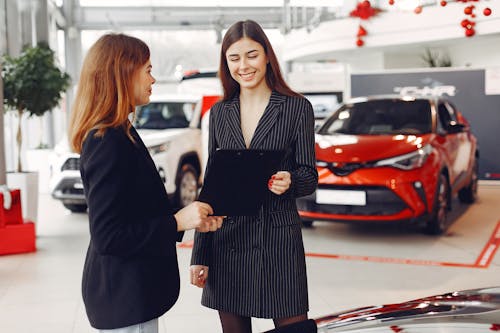 Cheerful young friendly dealer in formal stylish black dress showing contract to smiling female customer in black jacket while standing in car showroom against new shiny automobiles