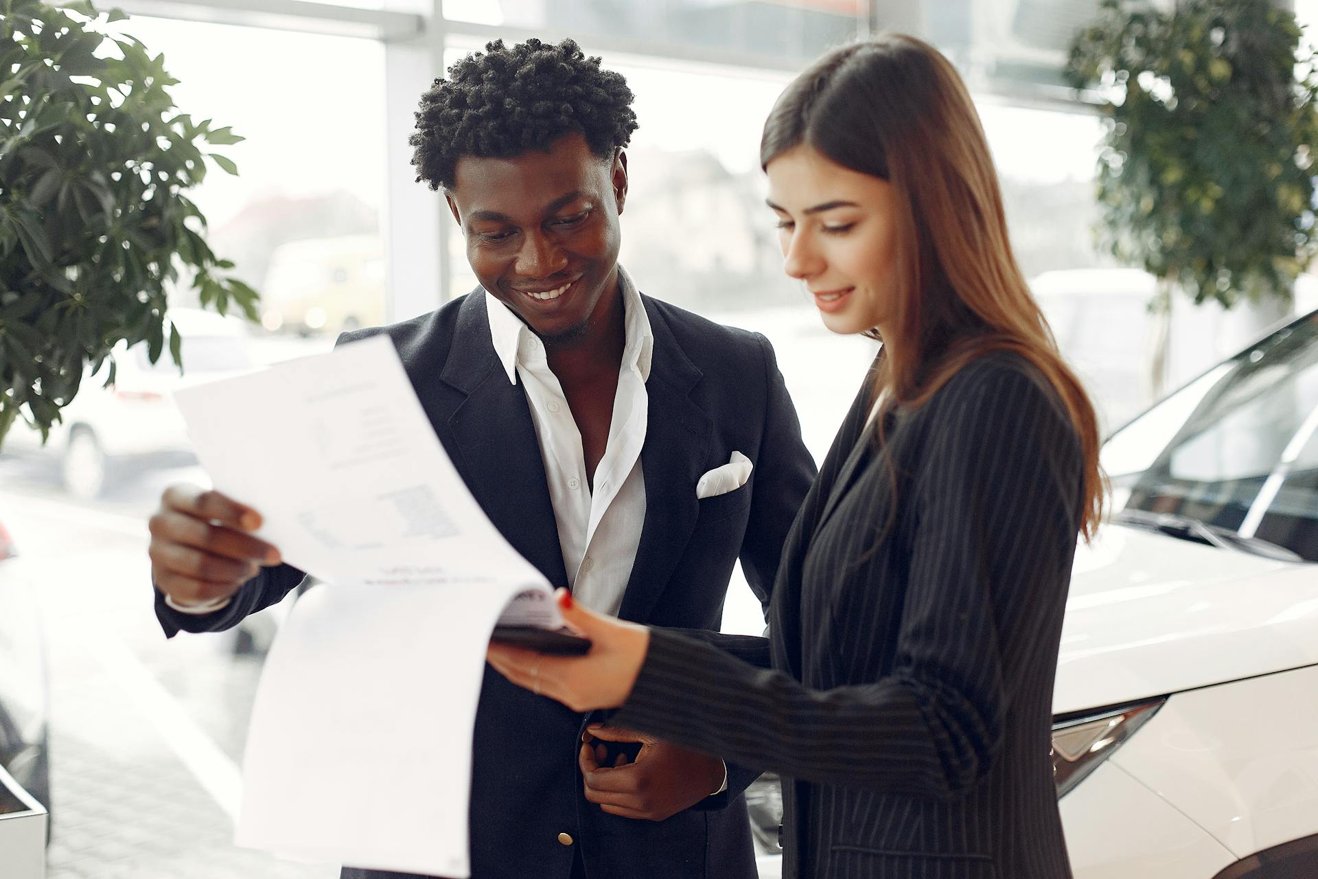 Cheerful smiling multiethnic businessman in classy suit and professional elegant female consultant standing close and reading contract details in car showroom