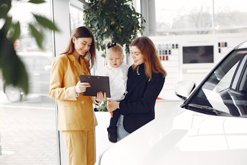 Positive young female with cute little daughter on hands discussing car characteristics with professional dealer in stylish beige suit while standing in car showroom in daylight