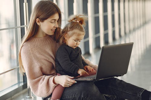 Free Cheerful young mother in casual clothes with cute blond girl on laps browsing netbook while sitting in modern building hallway in daylight and smiling Stock Photo