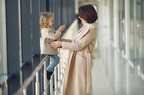 Free Side view of amazed woman in trendy beige coat standing near adorable funny daughter sitting on hallway railing and clapping hands in daylight Stock Photo