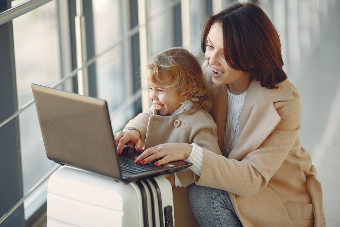 Cheerful stylish woman with suitcase hugging adorable laughing daughter and browsing laptop together while waiting for flight in airport hallway