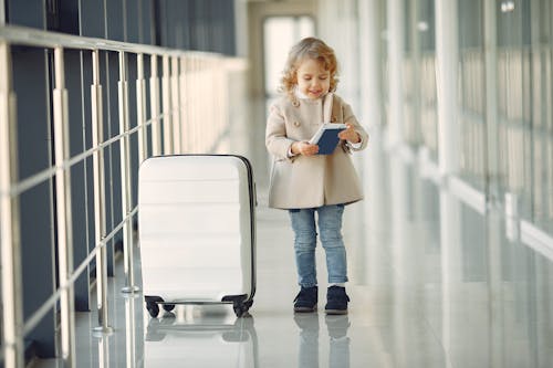 Free Full body of smiling cute little girl in jeans and beige coat standing near suitcase and checking information in documents Stock Photo