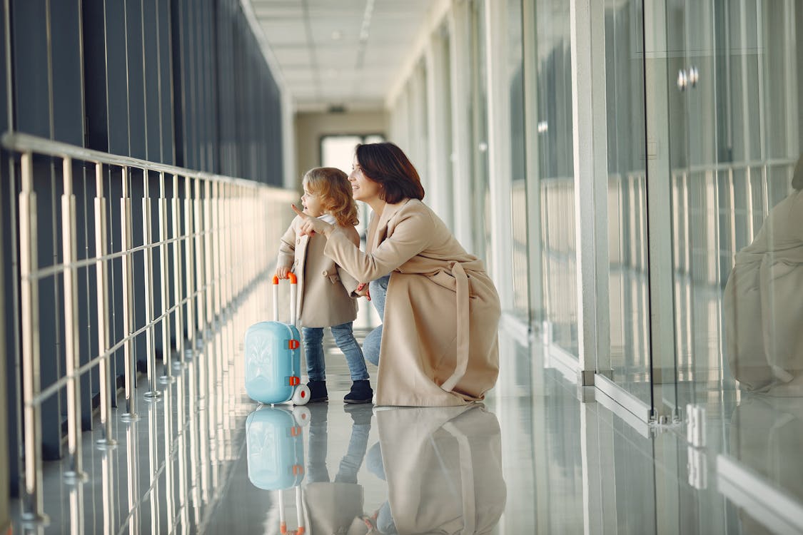 Free Side view full body of astonished cute little girl with kids suitcase and smiling mother pointing out window while walking together in contemporary airport hallway Stock Photo