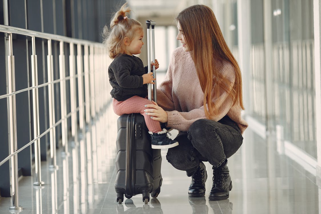 Free Full length of positive content woman and cute stylish blond little girl sitting on black suitcase in airport corridor while waiting for flight Stock Photo
