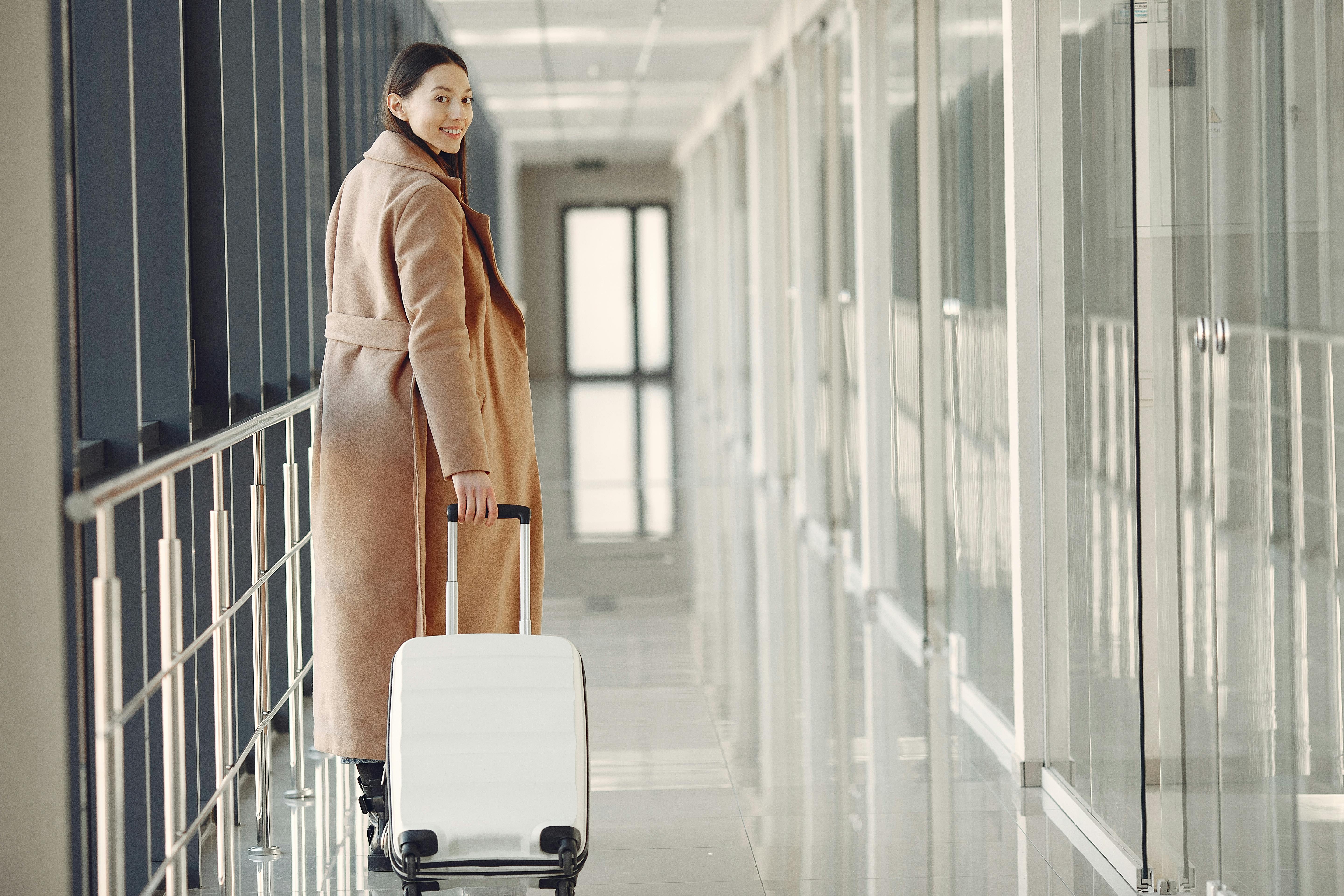 Woman in a beige coat pulls a white suitcase down a bright modern hallway.