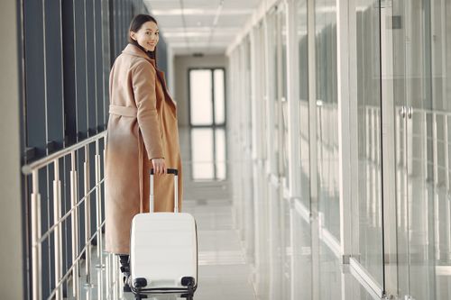 Free Back view full length of young cheerful female passenger in trendy beige coat walking along airport airbridge and looking at camera over shoulder Stock Photo