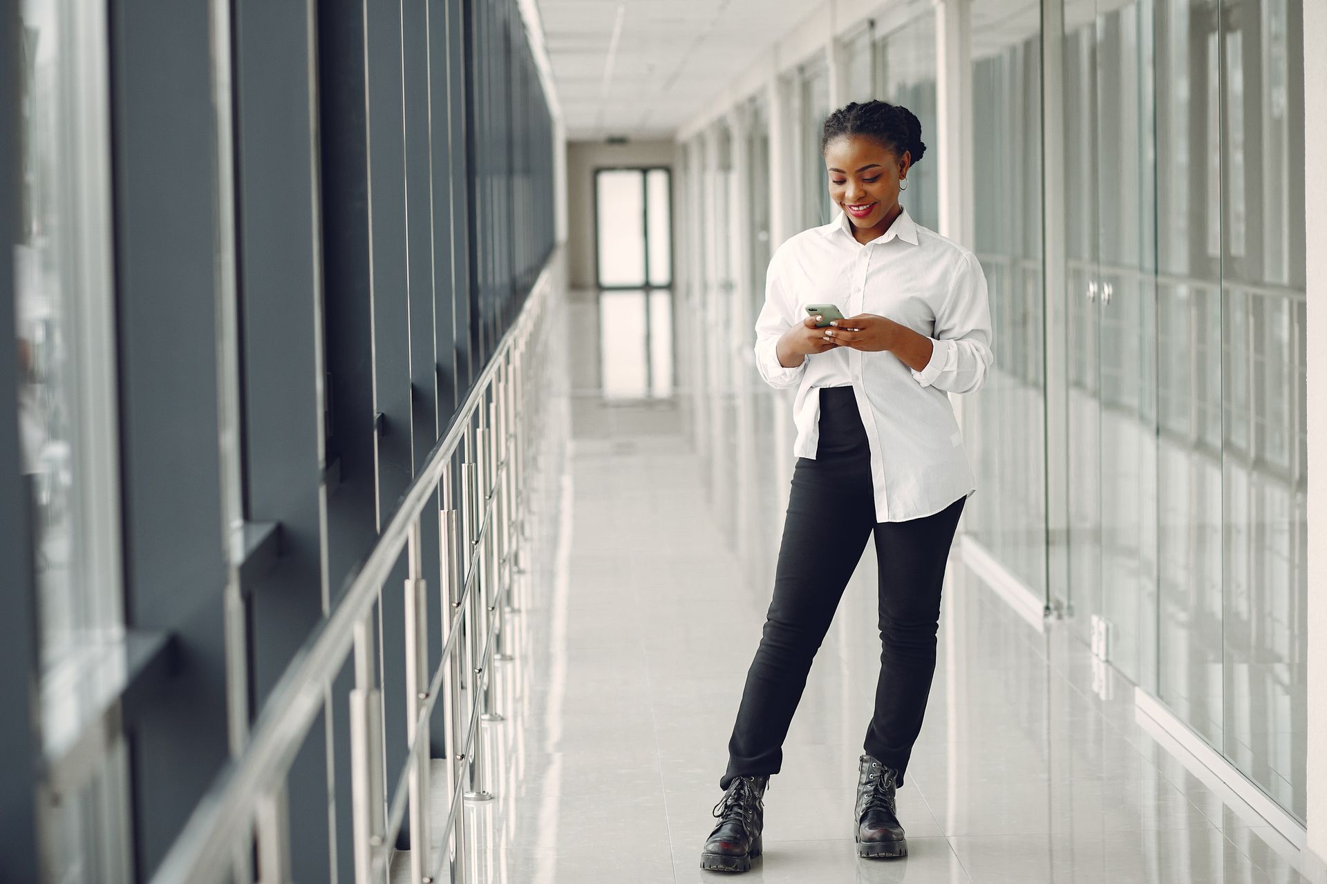 Full length of optimistic positive African American female office worker in formal clothes text messaging on cellphone while standing in modern office building