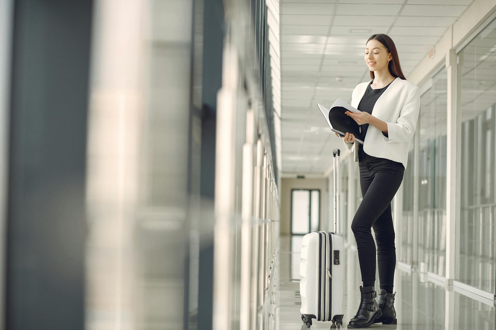Full body of slim female businesswoman in formal clothes with suitcase standing in airport hallway and reading note in notebook while waiting for flight
