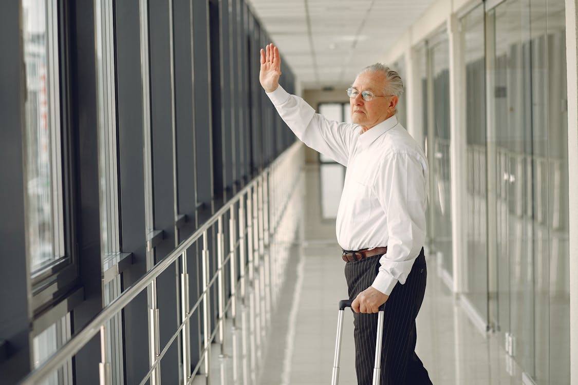 Free Side view of aged calm male in formal clothes with suitcase walking along modern airport hallway and waving goodbye while looking out glass wall Stock Photo