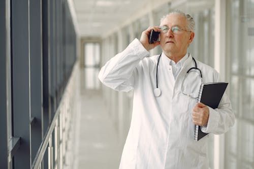 Serious mature male doctor in medical uniform standing with stethoscope and notebook while talking on smartphone in clinic corridor