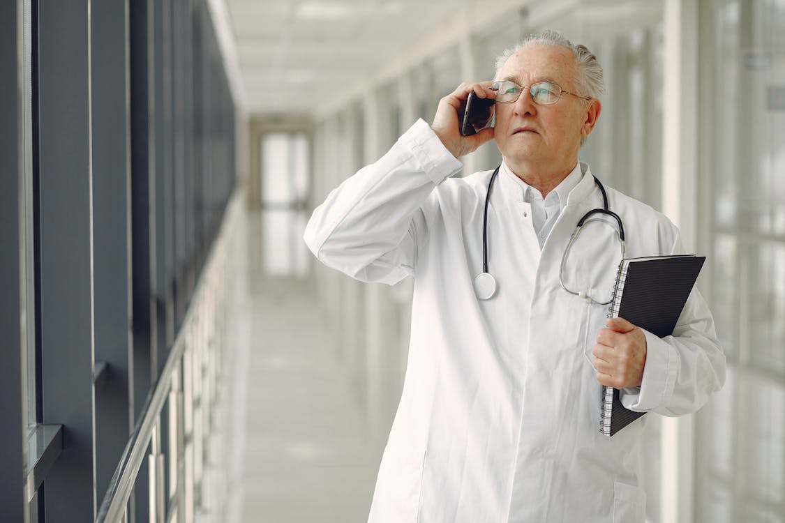 Free Serious mature male doctor in medical uniform standing with stethoscope and notebook while talking on smartphone in clinic corridor Stock Photo