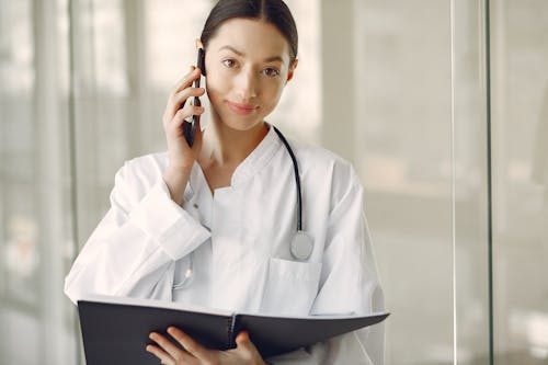 Free Positive doctor in medical uniform talking on cellphone in clinic corridor Stock Photo